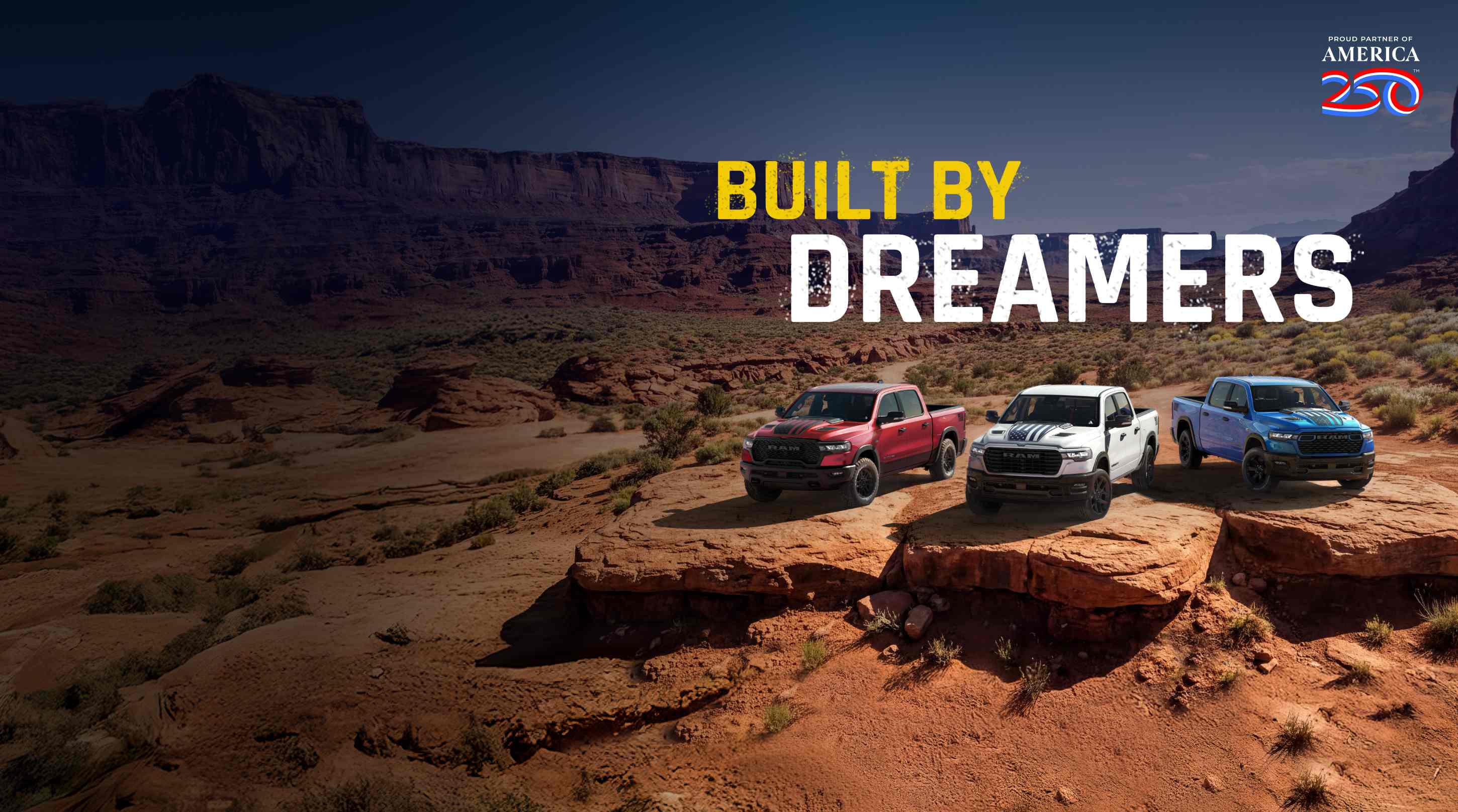 Three 2026 Ram trucks parked side-by-side on an outcropping in the desert. From left to right; a red Ram 1500 Rebel 4x4 Crew Cab, a white Ram 1500 Laramie 4x4 Crew Cab and a blue Ram 1500 Big Horn 4x4 Crew Cab. Proud Partner Of America 250. Built By Dreamers.
