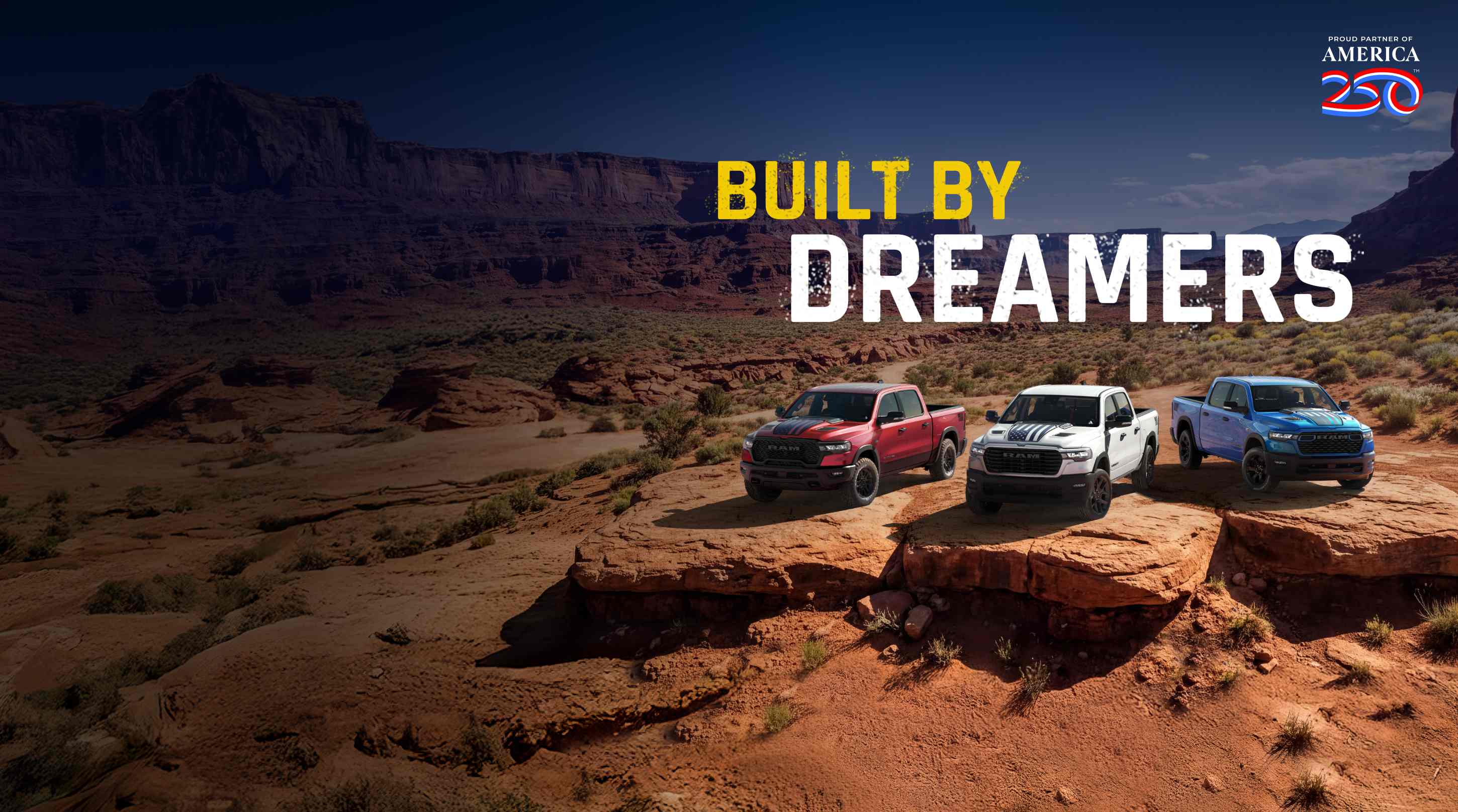 Three 2026 Ram trucks parked side-by-side on an outcropping in the desert. From left to right; a red Ram 1500 Rebel 4x4 Crew Cab, a white Ram 1500 Laramie 4x4 Crew Cab and a blue Ram 1500 Big Horn 4x4 Crew Cab. Proud Partner Of America 250. Built By Dreamers.