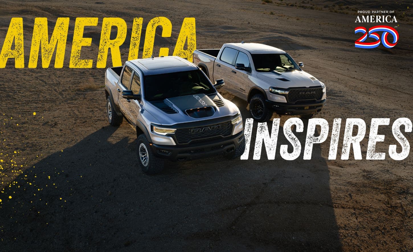 Two Ram Brand Off-Road Trucks parked off-road in the desert: on the left, a silver 2025 Ram 1500 RHO Crew Cab and on the right a silver 2025 Ram 1500 Rebel X Crew Cab. Proud partner of America 250.