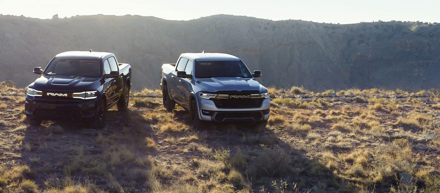 Two Ram Brand electric vehicles parked on a grassy hilltop. On the left a 2025 Ram 1500 Rev Tungsten 4x4 Crew Cab and on the right, a 2025 Ram 1500 Ramcharger Limited 4x4 Crew Cab.