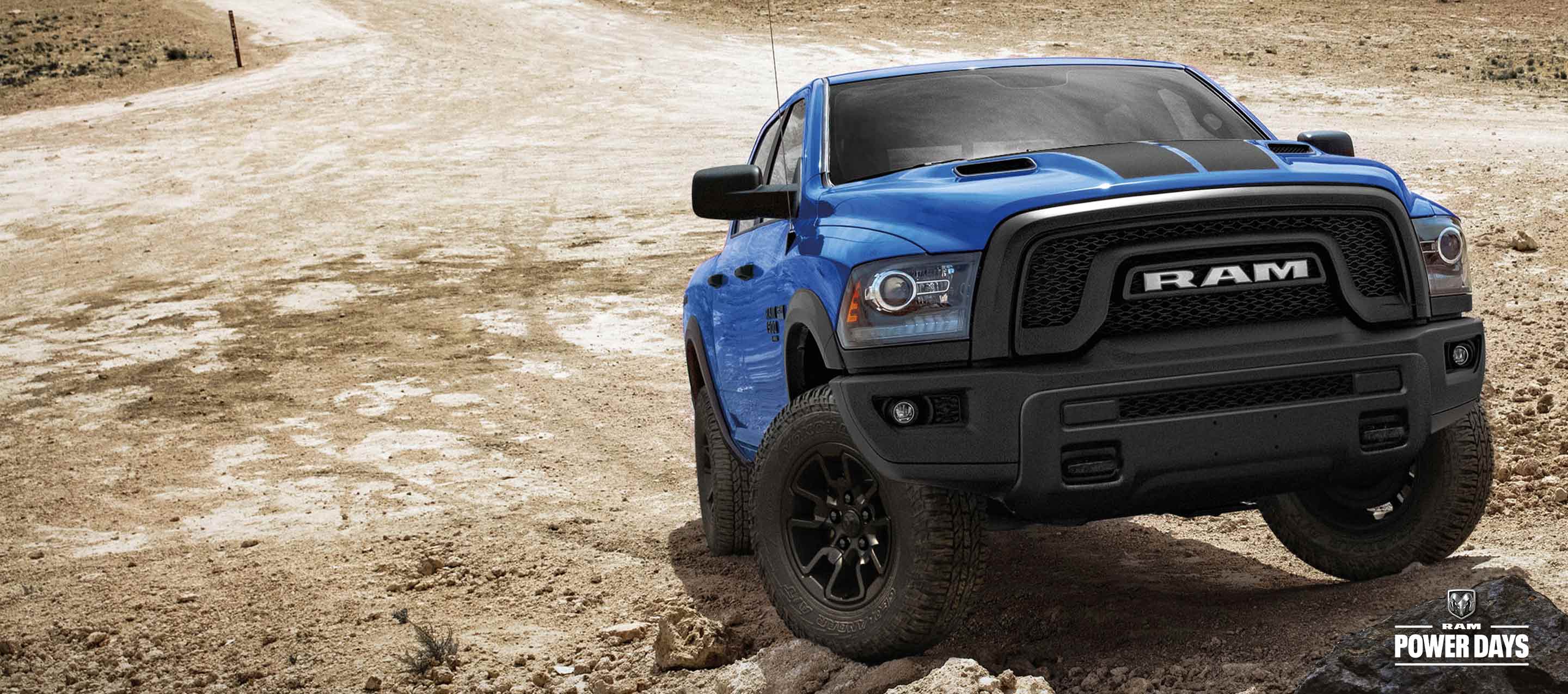 A blue 2023 Ram 1500 Classic Warlock Crew Cab with the All Terrain Package climbing a sandy hill off-road.