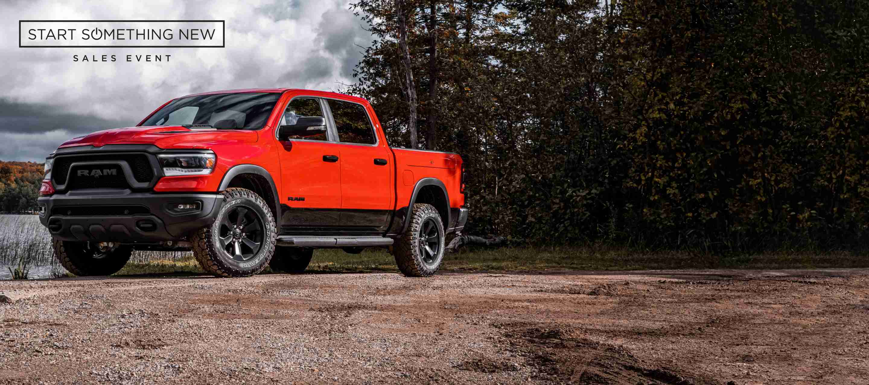 A red 2023 Ram 1500 Rebel 4x4 Crew Cab parked beside a lake, with storm clouds overhead. Start of Something New Sales Event.