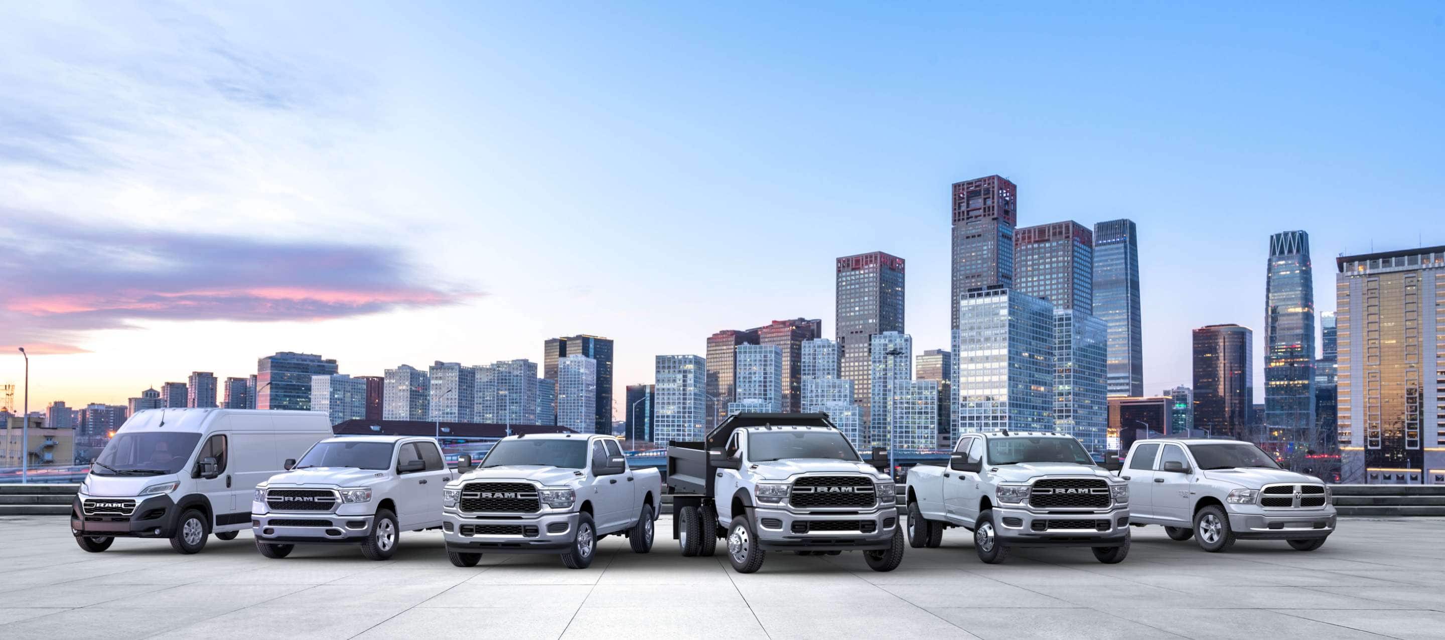 The Ram Brand lineup, all in white, parked side-by-side with a cityscape at dusk in the background. Beginning on the left: a 2023 Ram ProMaster 3500 High Roof Cargo Van, 2022 Ram ProMaster City Cargo Van, 2023 Ram 1500 Tradesman Crew Cab, 2023 Ram 5500 Tradesman Regular Cab with dump body, 2023 Ram 3500 Tradesman Crew Cab and 2023 Ram 1500 Classic Regular Cab. Ram Commercial.
