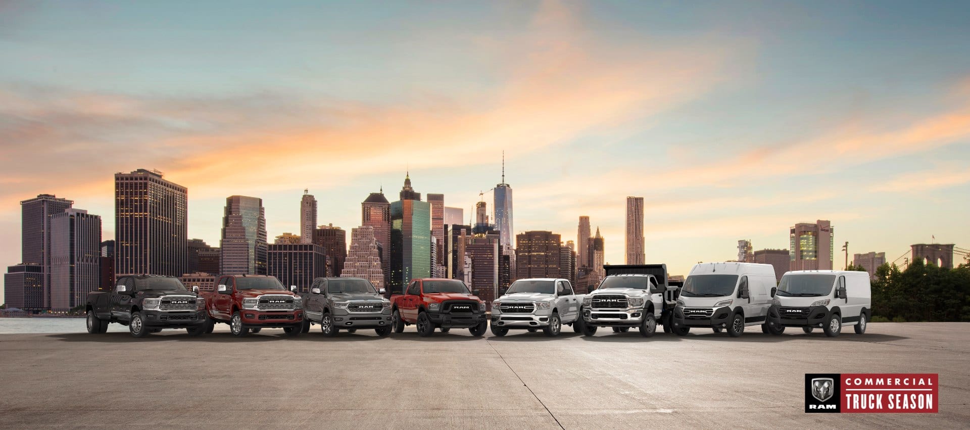 The Ram Brand lineup parked side-by-side at dusk, with a cityscape in the background. From left to right: a black 2024 Ram 3500 Limited Crew Cab, a red 2024 Ram 2500 Limited Crew Cab, a gray 2024 Ram 1500 Limited Crew Cab, a red 2024 Ram 1500 Classic Warlock Crew Cab, a white 2024 Ram 1500 Tradesman Crew Cab, a white Ram 5500 Tradesman Chassis Regular Cab with dump body upfit, a white 2024 Ram ProMaster 2500 SLT High Roof Cargo Van and a white 2024 Ram ProMaster 1500 Tradesman Standard Roof Cargo Van. Ram Commercial Truck Season.