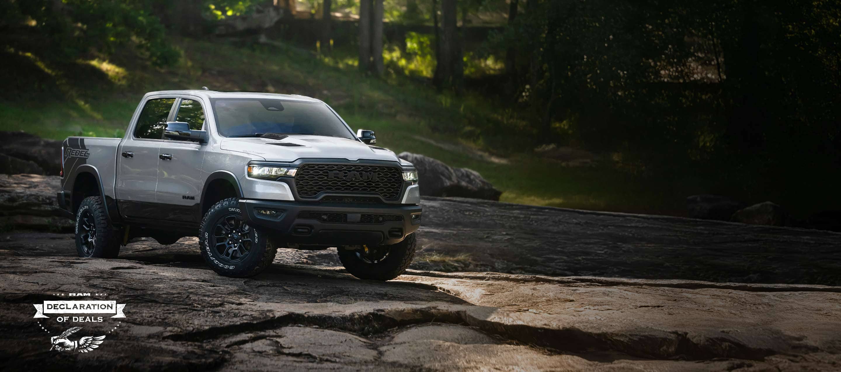 A white 2026 Ram 1500 Rebel 4x4 Crew Cab traveling down a rocky trail off-road, with a forest in the background. Declaration of Deals Sales Event.