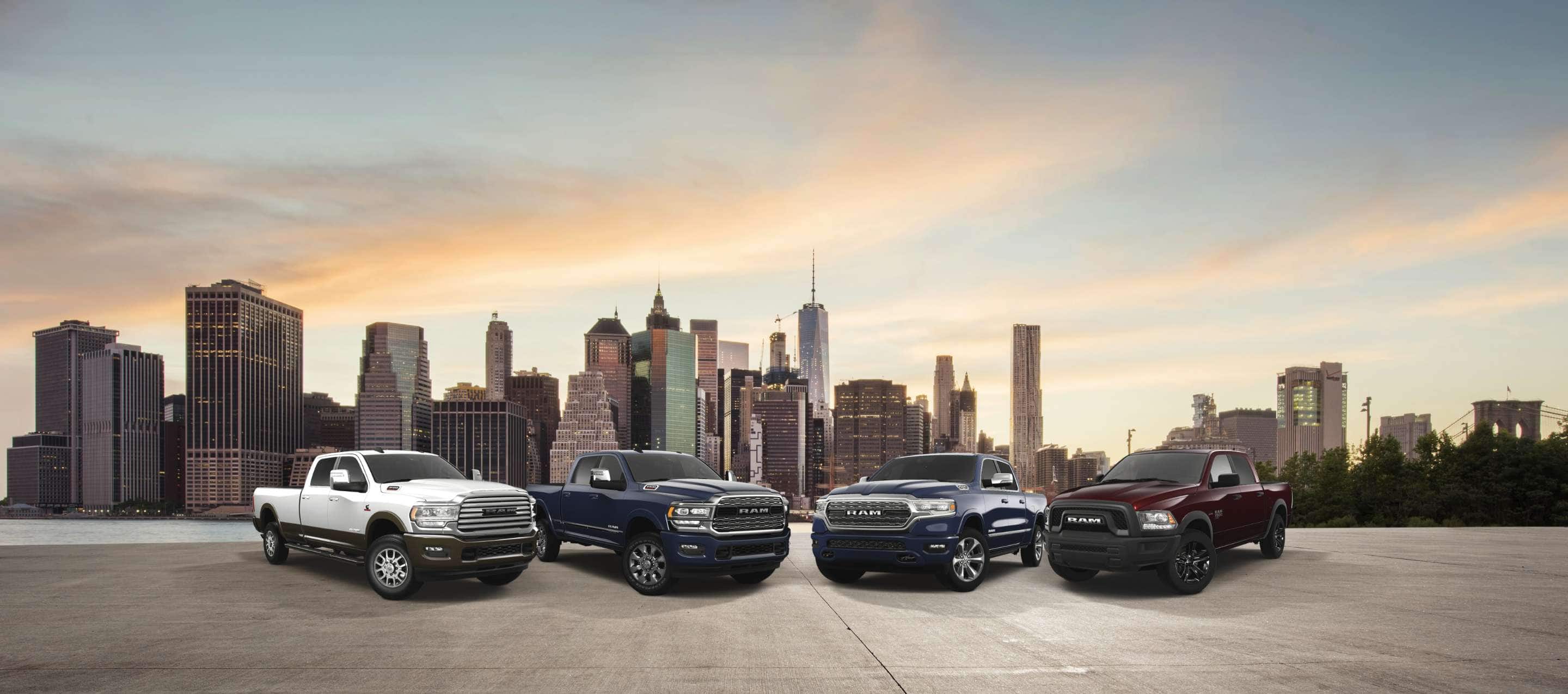 Four Ram Brand trucks parked on the waterfront, with a cityscape of high-rise buildings across the river. From left to right: a white 2023 Ram 3500 Limited Longhorn 4x4 Crew Cab, a blue 2023 Ram 2500 Limited 4x4 Crew Cab, a blue 2023 Ram 1500 Limited 4x4 Crew Cab and a red 2023 Ram 1500 Classic Warlock 4x4 Crew Cab.