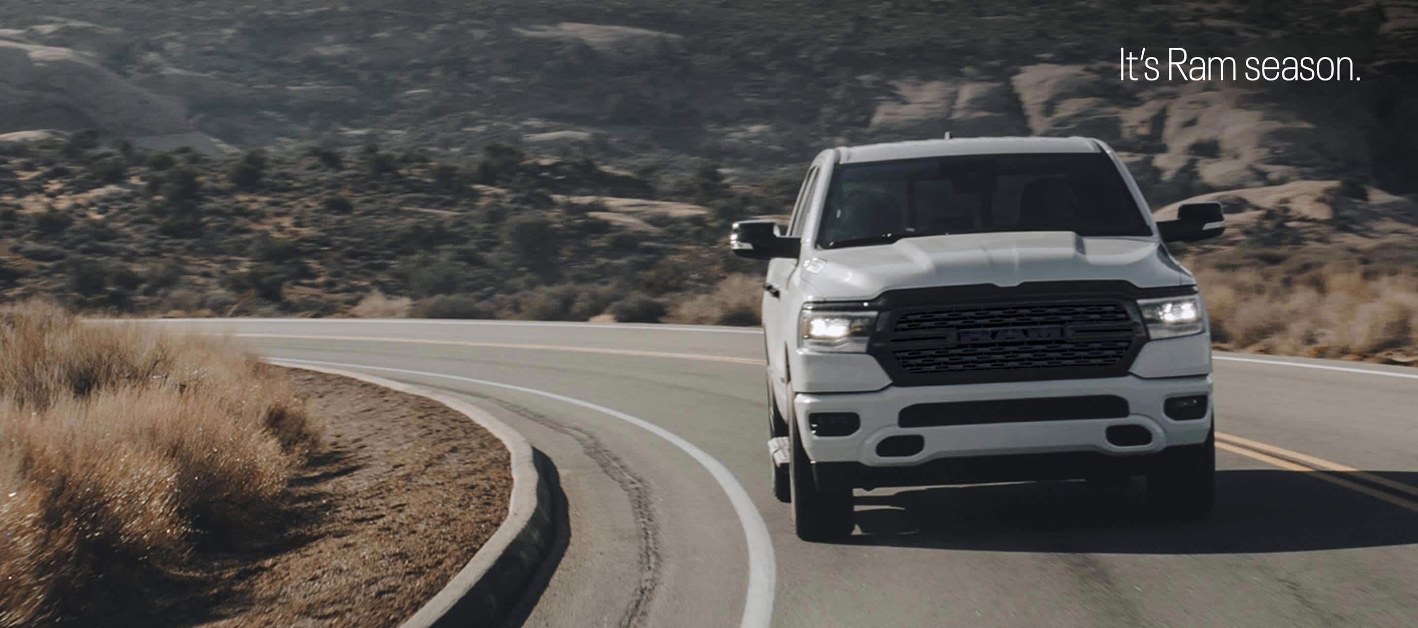 A head-on angle of a white 2023 Ram 1500 Big Horn 4x4 Crew Cab being driven up a curved mountain road in the desert. It's Ram Season.