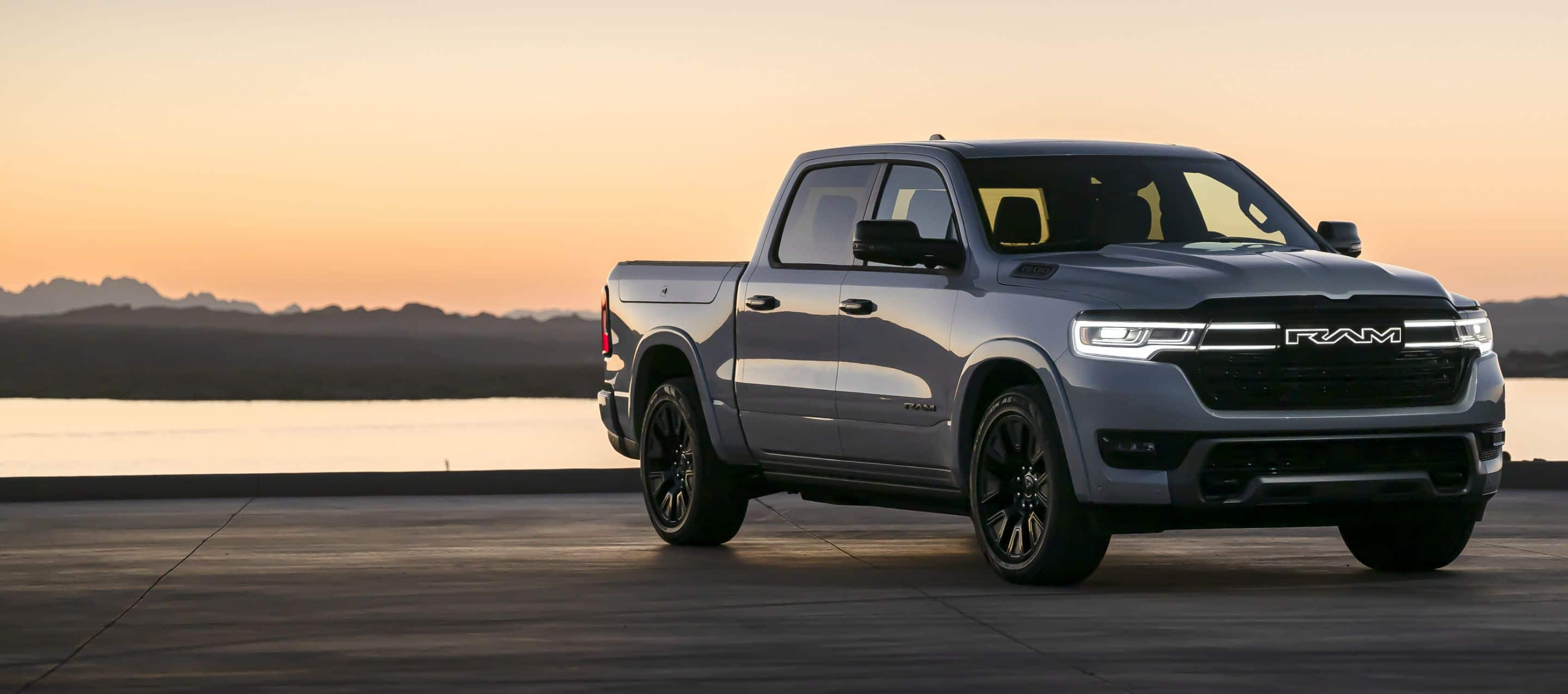 An angled passenger-side front profile of a gray 2025 Ram 1500 Ramcharger Limited Crew Cab with its headlamps and front end lighting on, parked near a lake at dusk.