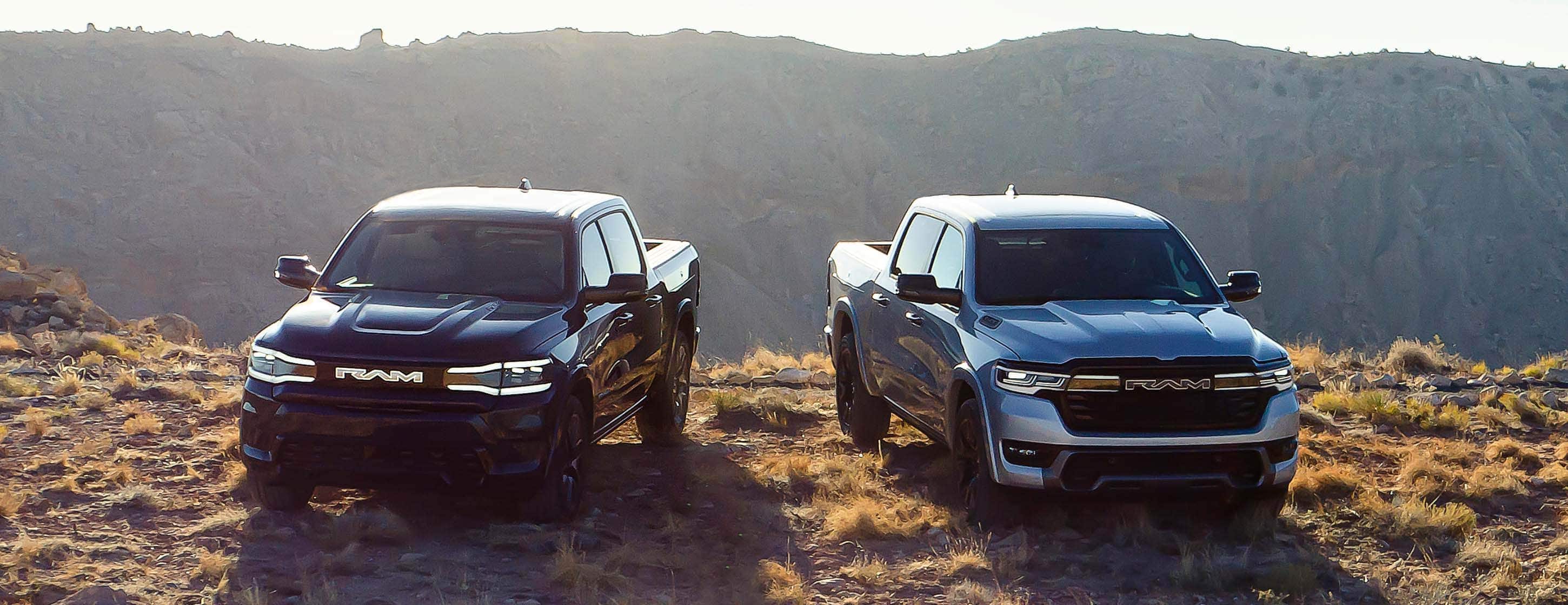 Two Ram Brand electric vehicles parked side by side on a grassy hilltop at sunrise with mountains in the distance. On the left a black 2025 Ram 1500 Rev Tungsten 4x4 Crew Cab and on the right, a blue 2025 Ram 1500 Ramcharger Limited 4x4 Crew Cab.