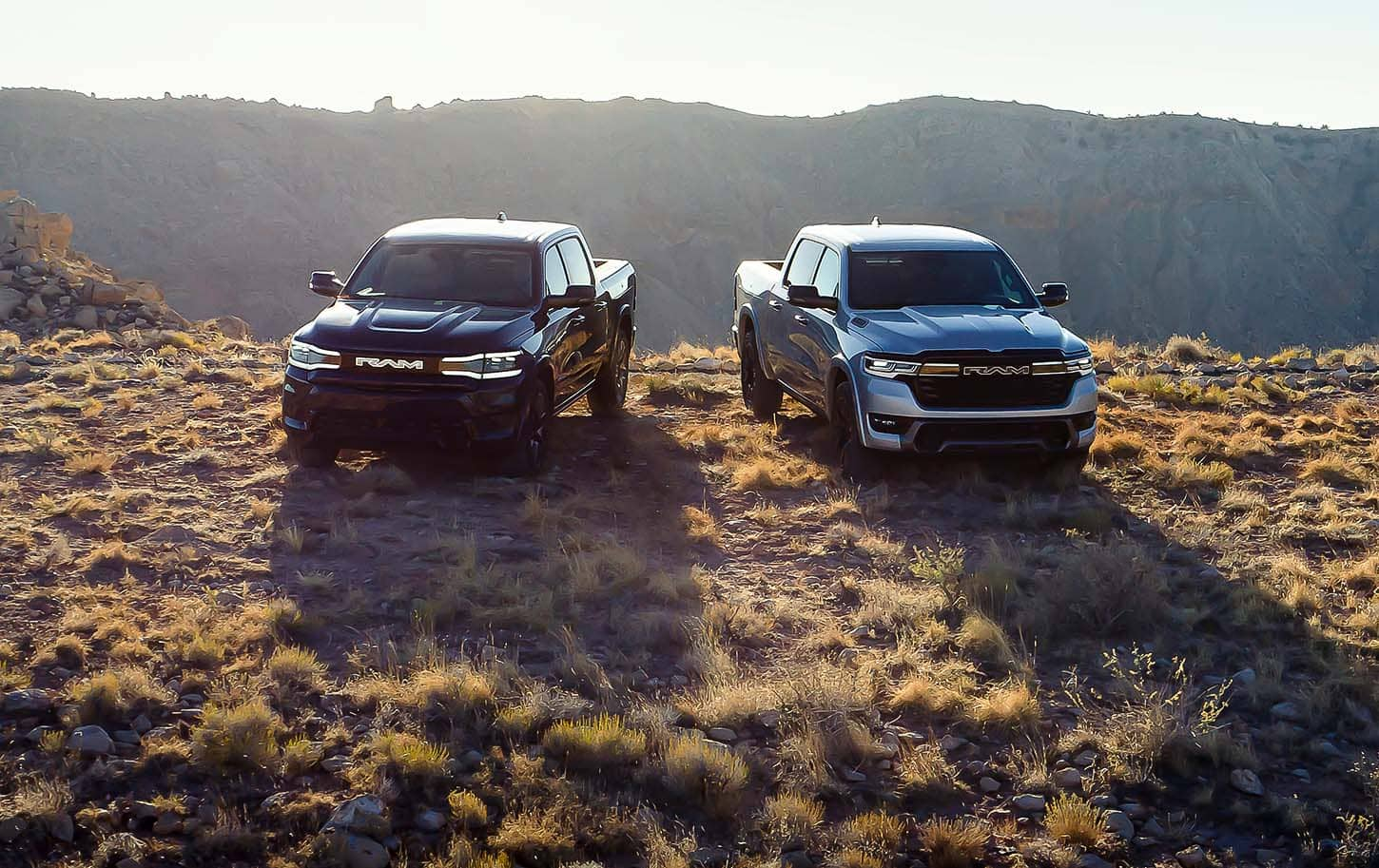 A gray 2025 Ram 1500 Rev Tungsten Crew Cab and a silver 2025 Ram 1500 Ramcharger Limited Crew Cab parked on a clearing in the mountains.