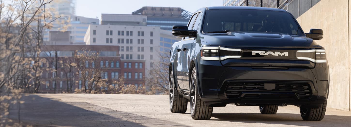 A blue 2025 Ram 1500 Rev Tungsten Crew Cab parked beside a freeway bridge with tall buildings in the background.