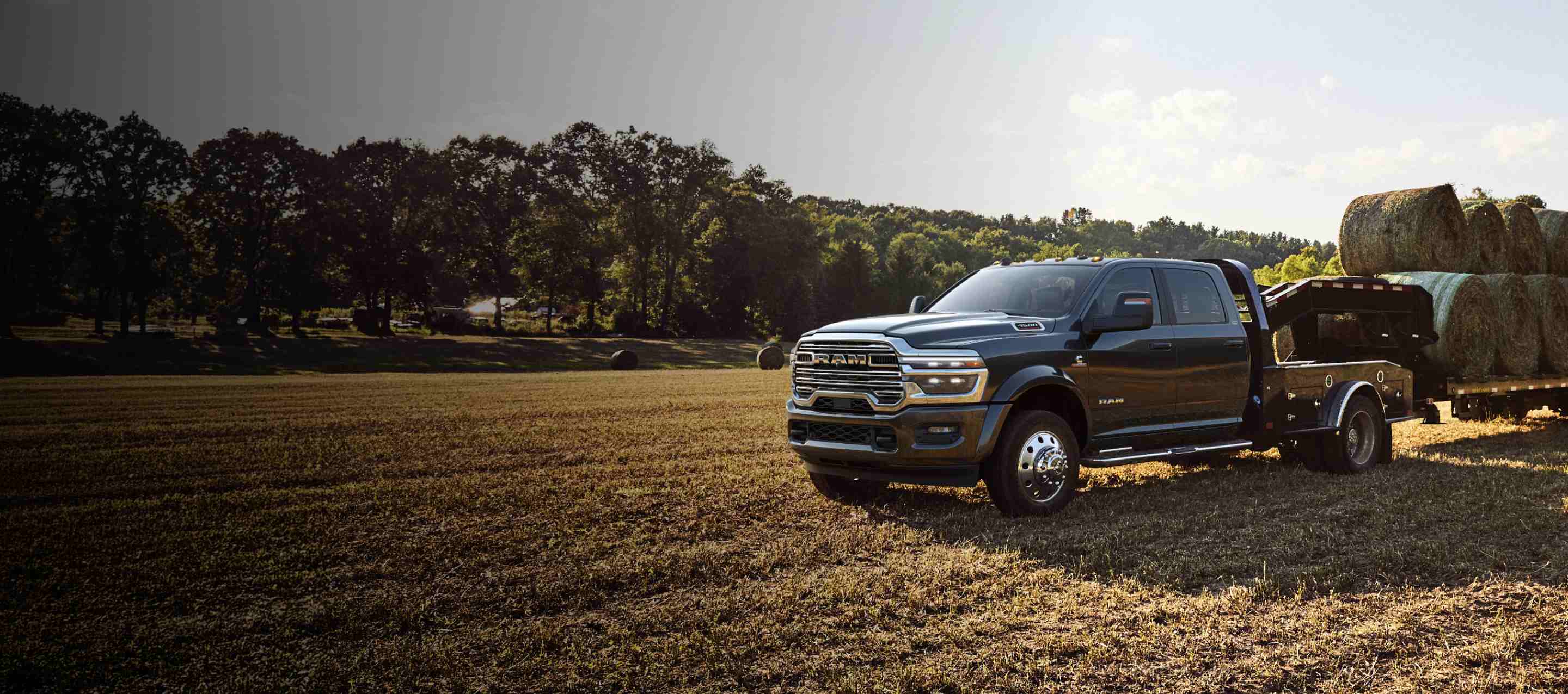A black 2026 Ram 4500 Big Horn Chassis Cab Crew Cab with the Laramie Trim Group and a gooseneck trailer loaded with several large bales of hay, parked in an open field.