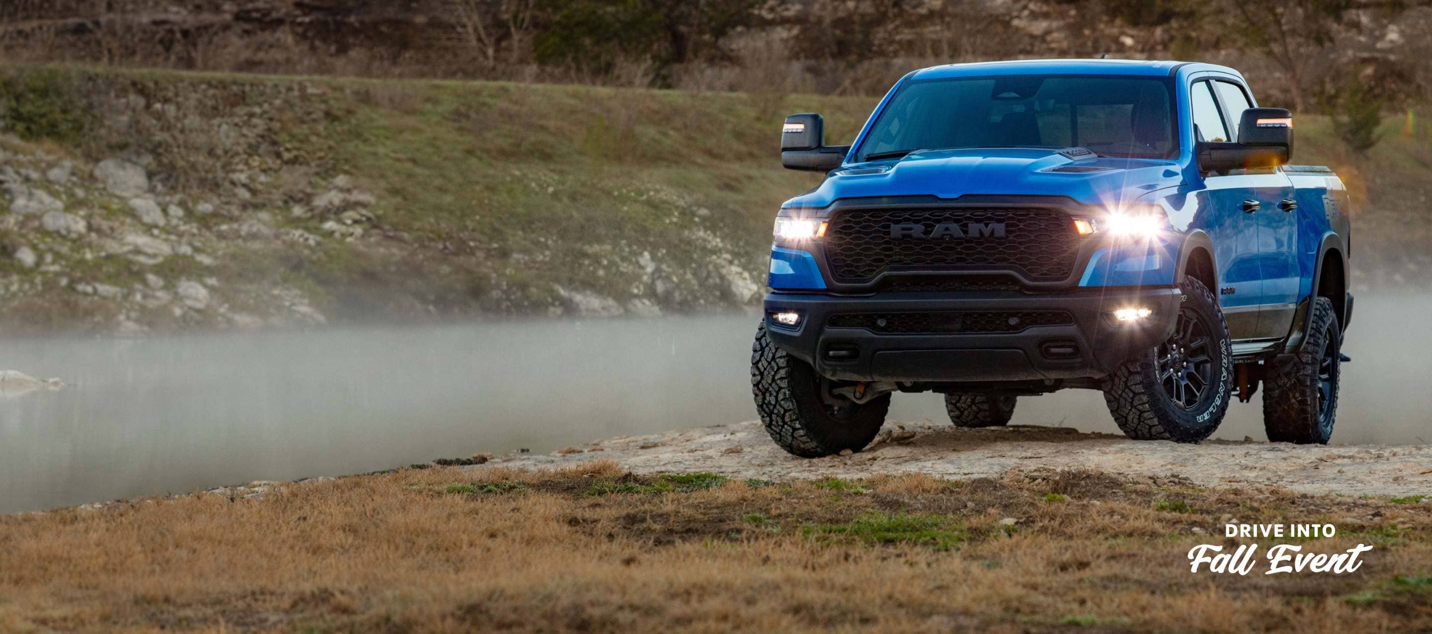 A blue 2025 Ram 1500 Rebel 4x4 Crew Cab with its headlamps and fog lamps on, traveling up a small hill off-road. Drive Into Fall Sales Event.
