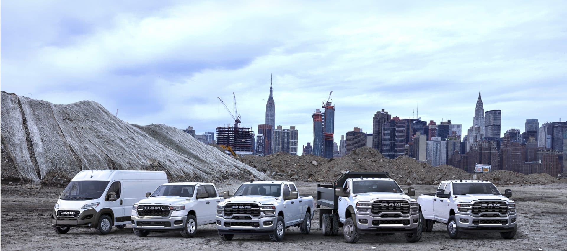 The 2025 Ram Brand lineup all in white, parked side-by-side at a pre-construction site, with a cityscape in the background. From left to right: a Ram ProMaster 3500 SLT High Roof Cargo Van, a Ram 1500 Tradesman 4x4 Crew Cab, a Ram 2500 Tradesman 4x4 Crew Cab, a Ram 5500 Tradesman Chassis Regular Cab with dump body upfit and a Ram 3500 Tradesman 4x4 Crew Cab.