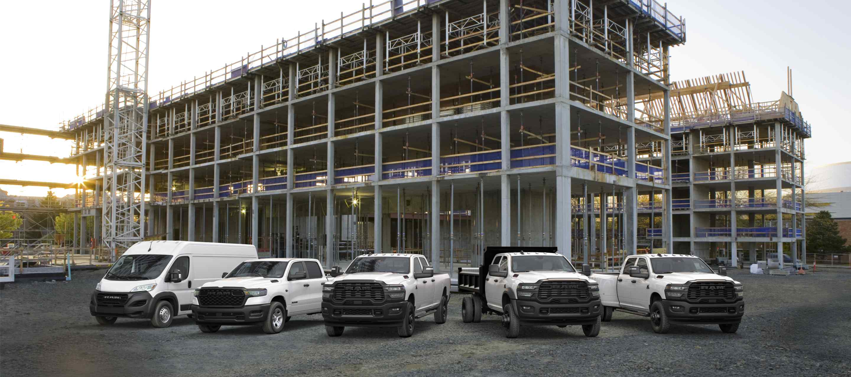 A lineup of 2026 Ram trucks and vans, all in white at a commercial construction site. From left to right: a Ram ProMaster 3500 Tradesman High Roof Cargo Van, a Ram 1500 Tradesman 4x4 Crew Cab, a Ram 2500 Tradesman 4x4 Crew Cab, a Ram 3500 Tradesman Crew Cab Chassis Cab with a dump body upfit and a Ram 3500 Tradesman 4x4 Crew Cab.