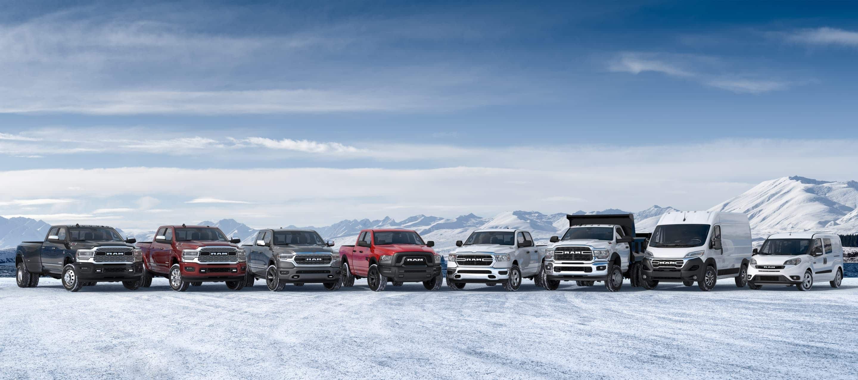 A lineup of six 2022 Ram trucks, a 2023 Ram ProMaster Cargo Van and a 2022 Ram ProMaster City Passenger Van, all parked on a snow-covered clearing near the water's edge of a mountain lake.