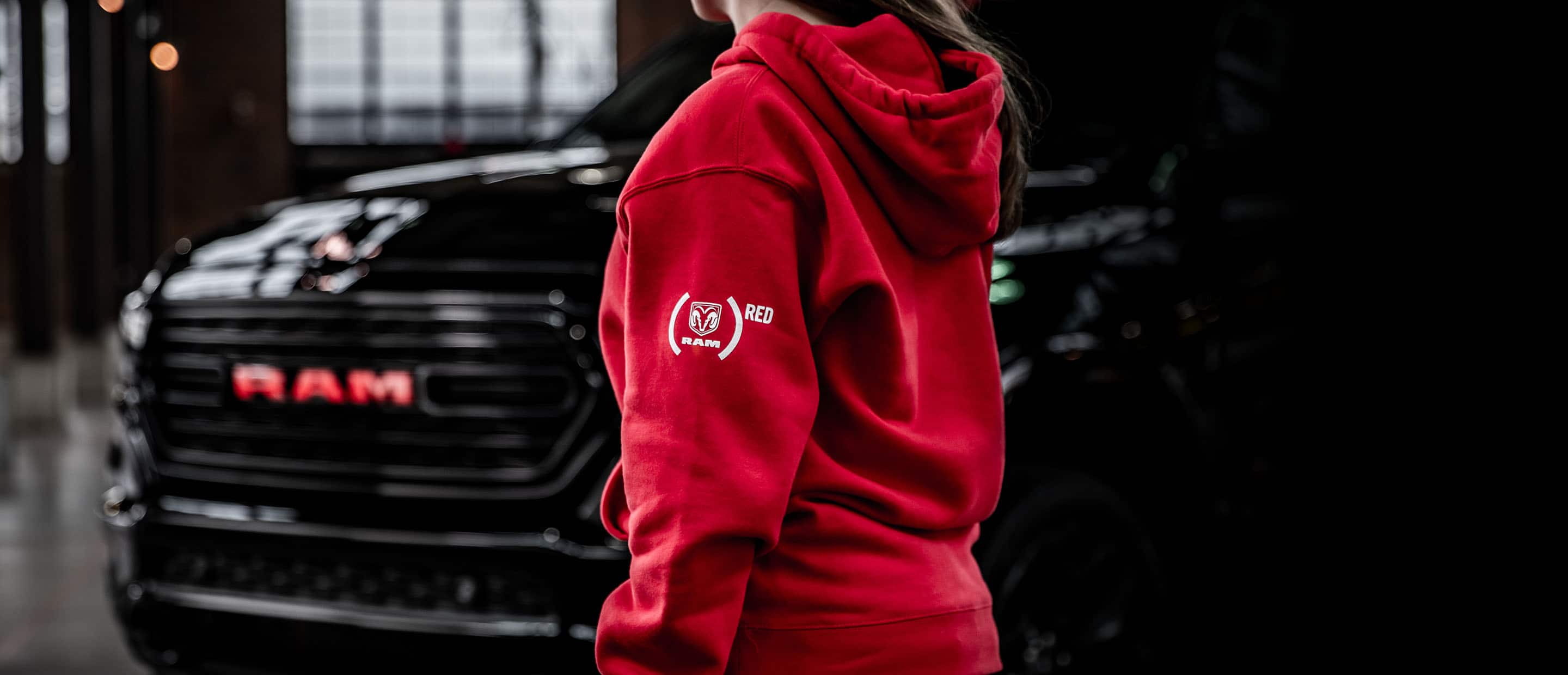 A woman wearing a red hoodie with  the Ram Red logo, walking toward a 2023 Ram 1500 Limited Ram Red Edition.