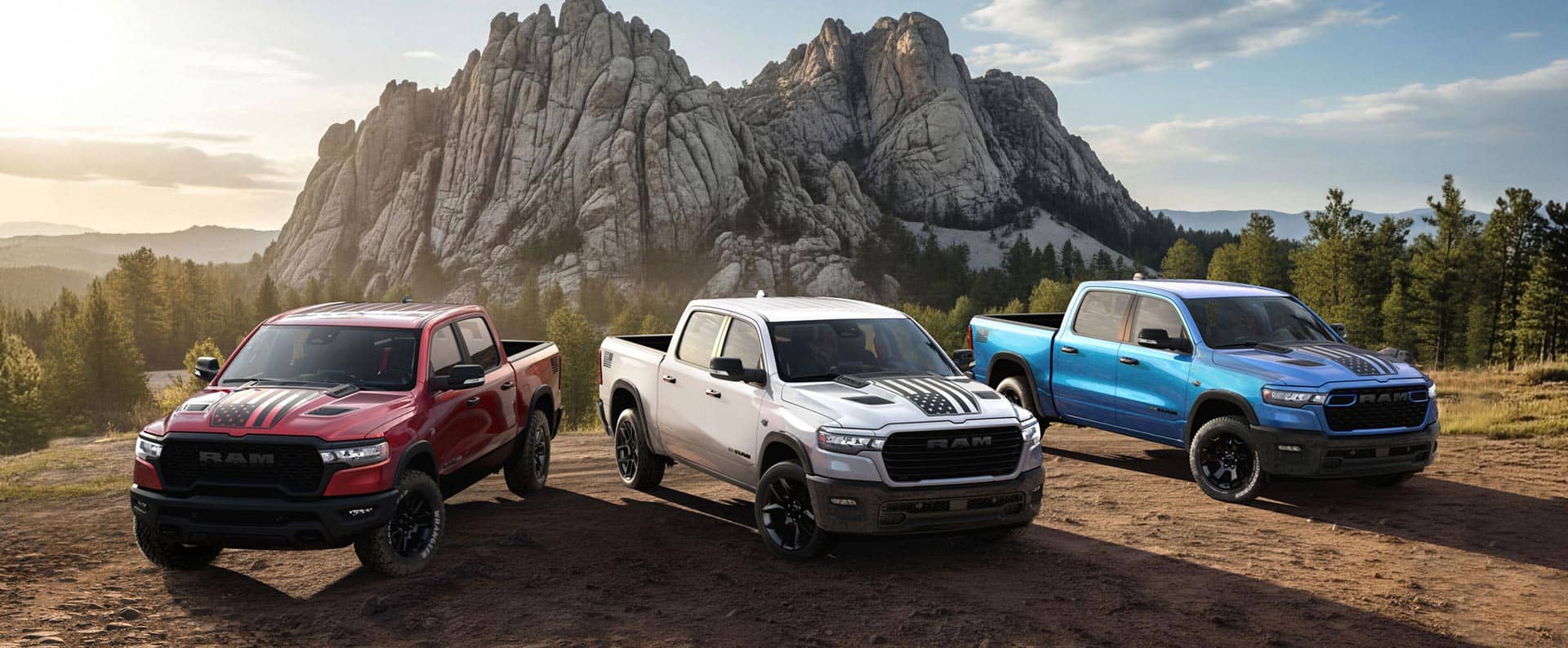 Three 2026 Ram 1500 America 250th Anniversary Edition pickups on a hilltop with mountains in the distance. From left to right; a red Ram 1500 Rebel 4x4 Crew Cab, a white Ram 1500 Laramie 4x4 Crew Cab and a blue Ram 1500 Big Horn 4x4 Crew Cab.