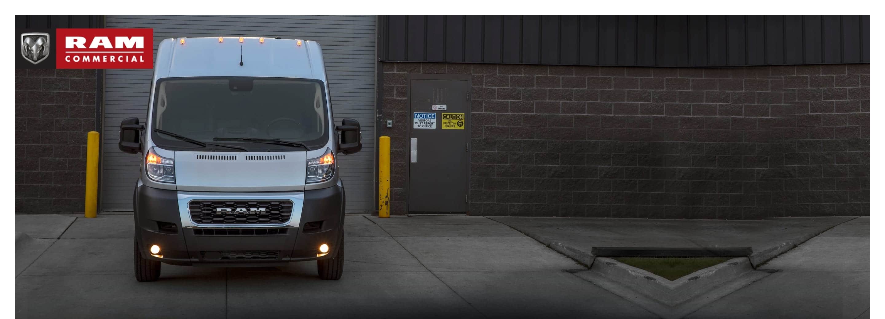Ram's head logo. Ram Commercial. A head-on view of the 2022 Ram ProMaster parked in front of a loading dock with its daytime running lamps on.