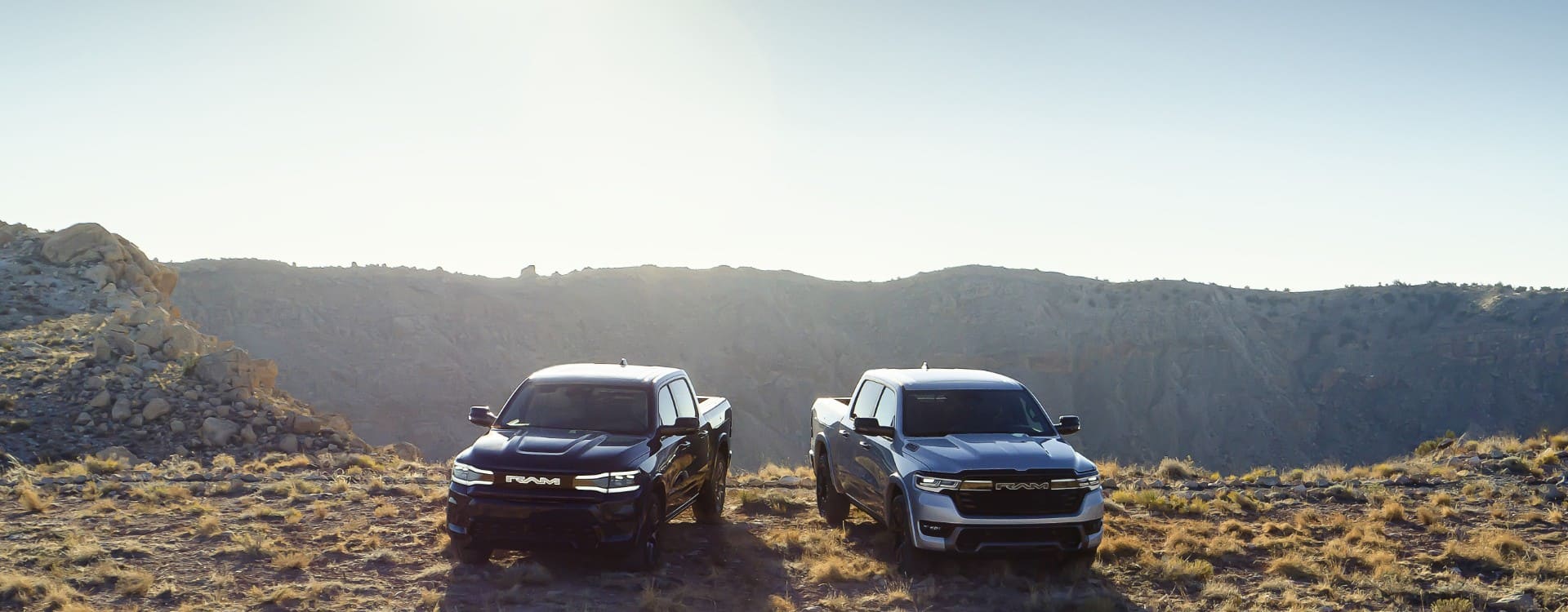 A 2025 Ram 1500 Rev Tungsten and a 2025 Ram 1500 Ramcharger Limited parked side-by-side on a grassy hilltop in the mountains.