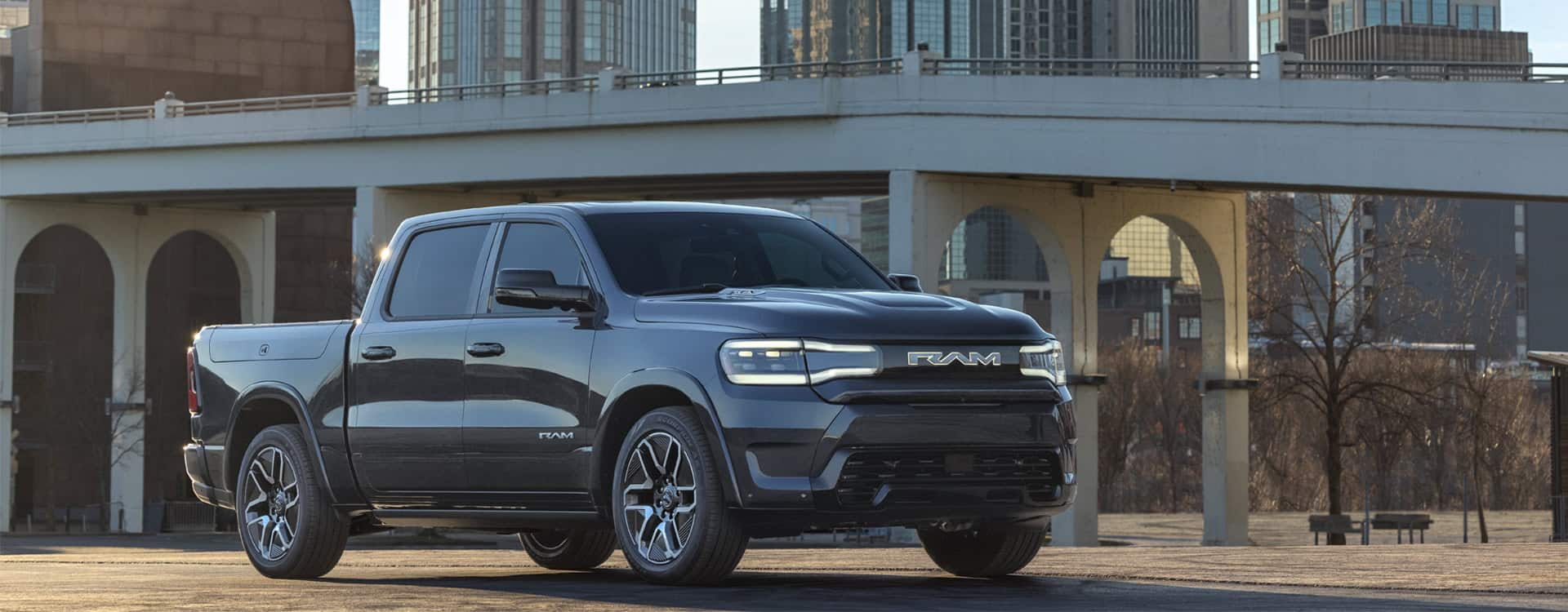 An angled passenger-side profile of a black 2025 Ram 1500 Rev Tungsten 4x4 Crew Cab parked in a city with a bridge and tall buildings in the background.