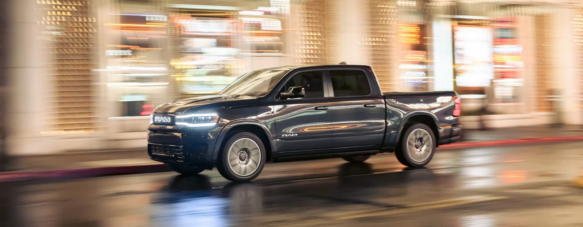 A gray 2025 Ram 1500 Rev Tungsten being driven at night on a city street bathed in lights. The background is blurred to indicate the vehicle is in motion.