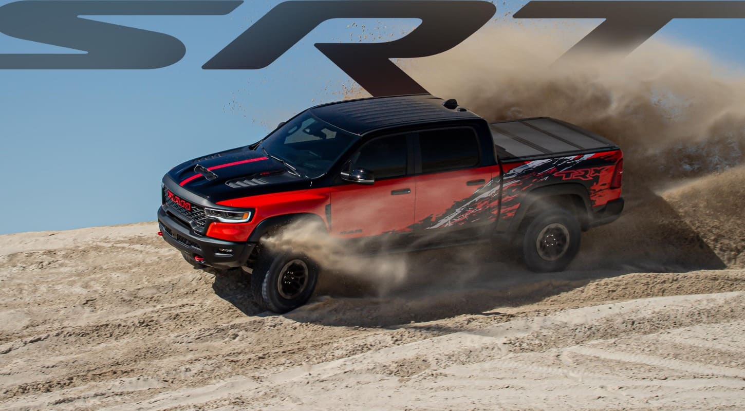 A red 2027 Ram 1500 TRX SRT Bloodshot Night Edition 4x4 Crew Cab with a black hood and roof and special bedside splash graphics, kicking up sand as it travels through the desert. SRT.