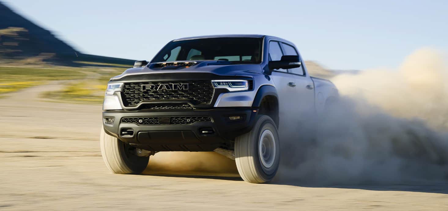 A silver 2025 Ram 1500 RHO Crew Cab traveling off-road in the desert, with dust and sand billowing from its tires.