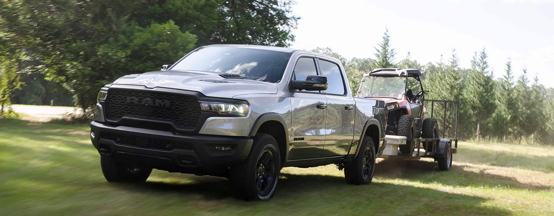 A silver 2024 Ram 2500 Rebel 4x4 Crew Cab being driven on a grassy trail off-road as it tows an ATV.