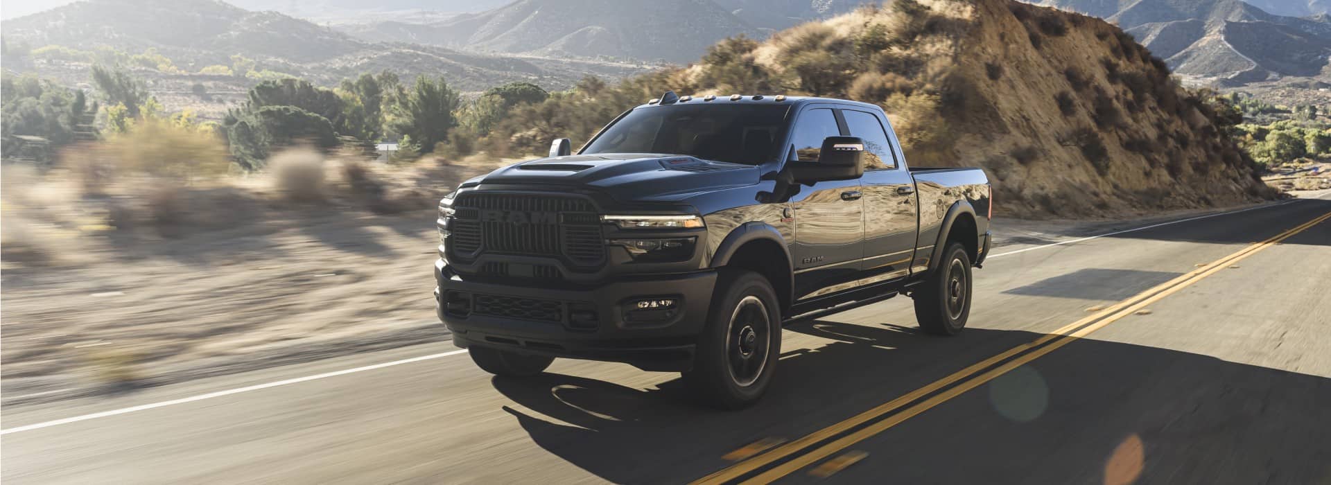 A black 2026 Ram 2500 Rebel Crew Cab traveling on a desert highway with mountains in the background.