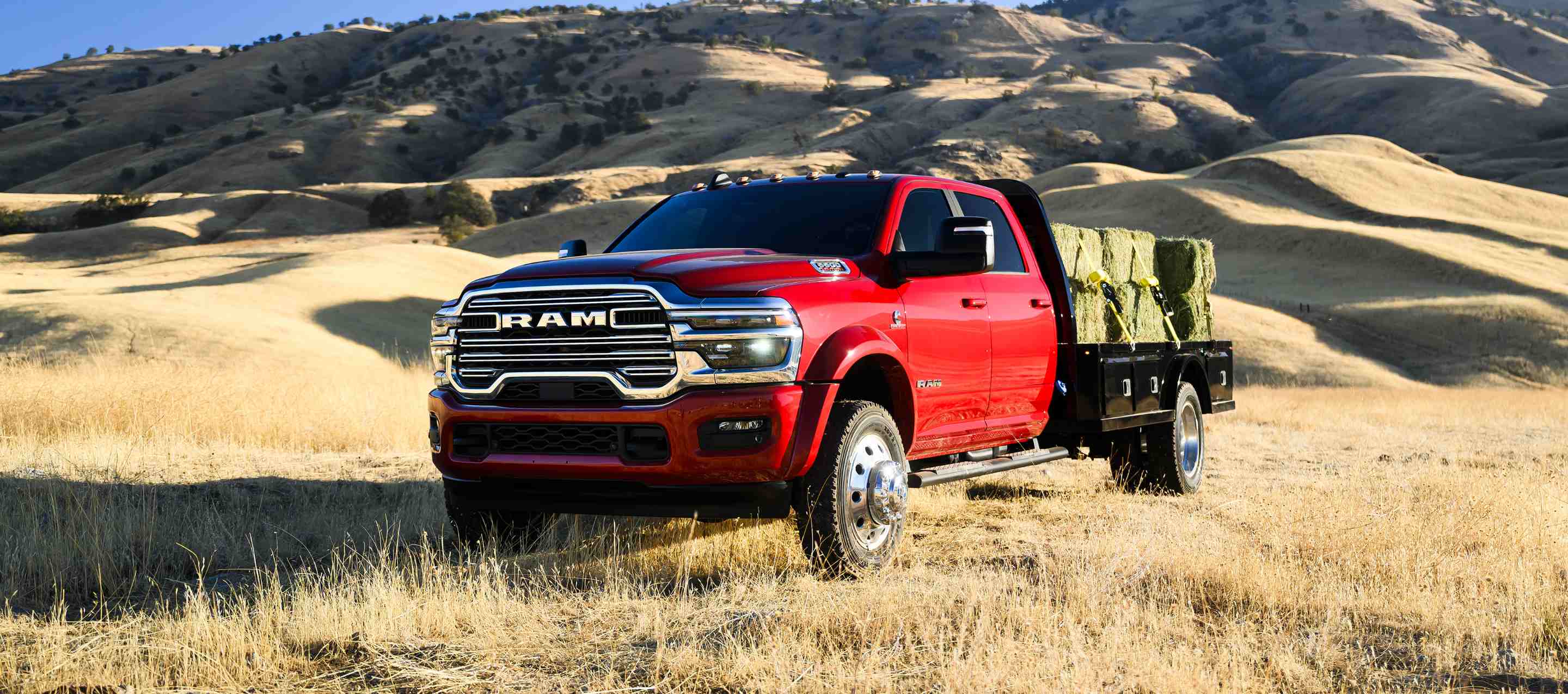A red 2025 Ram 5500 Laramie Chassis Cab Crew Cab carrying several large bales of hay on a long platform upfit.